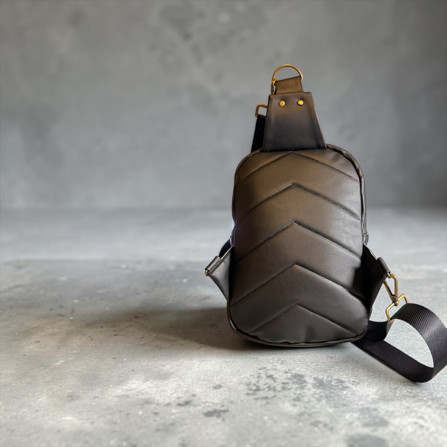 NspiVibe black leather crossbody sling bag with chevron-quilted back panel and brass-tone hardware on grey background